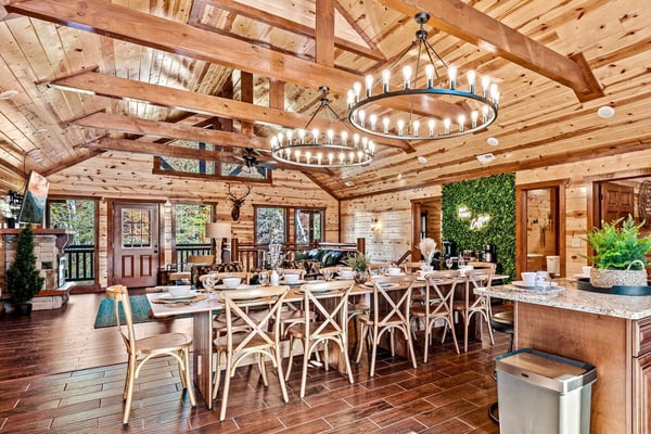 Spacious log cabin dining and kitchen area with exposed beam ceiling, circular chandeliers, long dining table with cross-back chairs, and granite countertops.