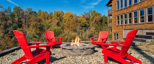 Four red Adirondack chairs surrounding a stone fire pit with burning flames, on gravel patio beside log cabin with autumn forest backdrop