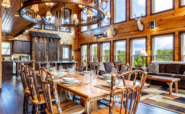 Rustic log cabin interior with dining table, birch-branch chairs, decorative chandelier, mountain views through large windows, and mounted deer heads.