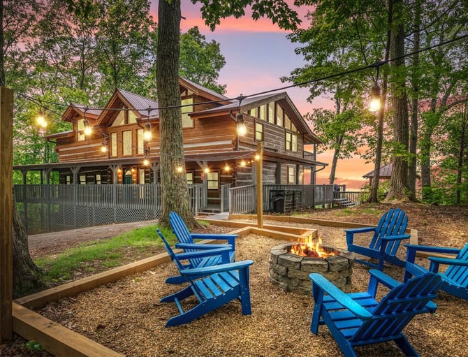 Log cabin with lit windows and blue Adirondack chairs around stone fire pit at dusk, surrounded by tall trees